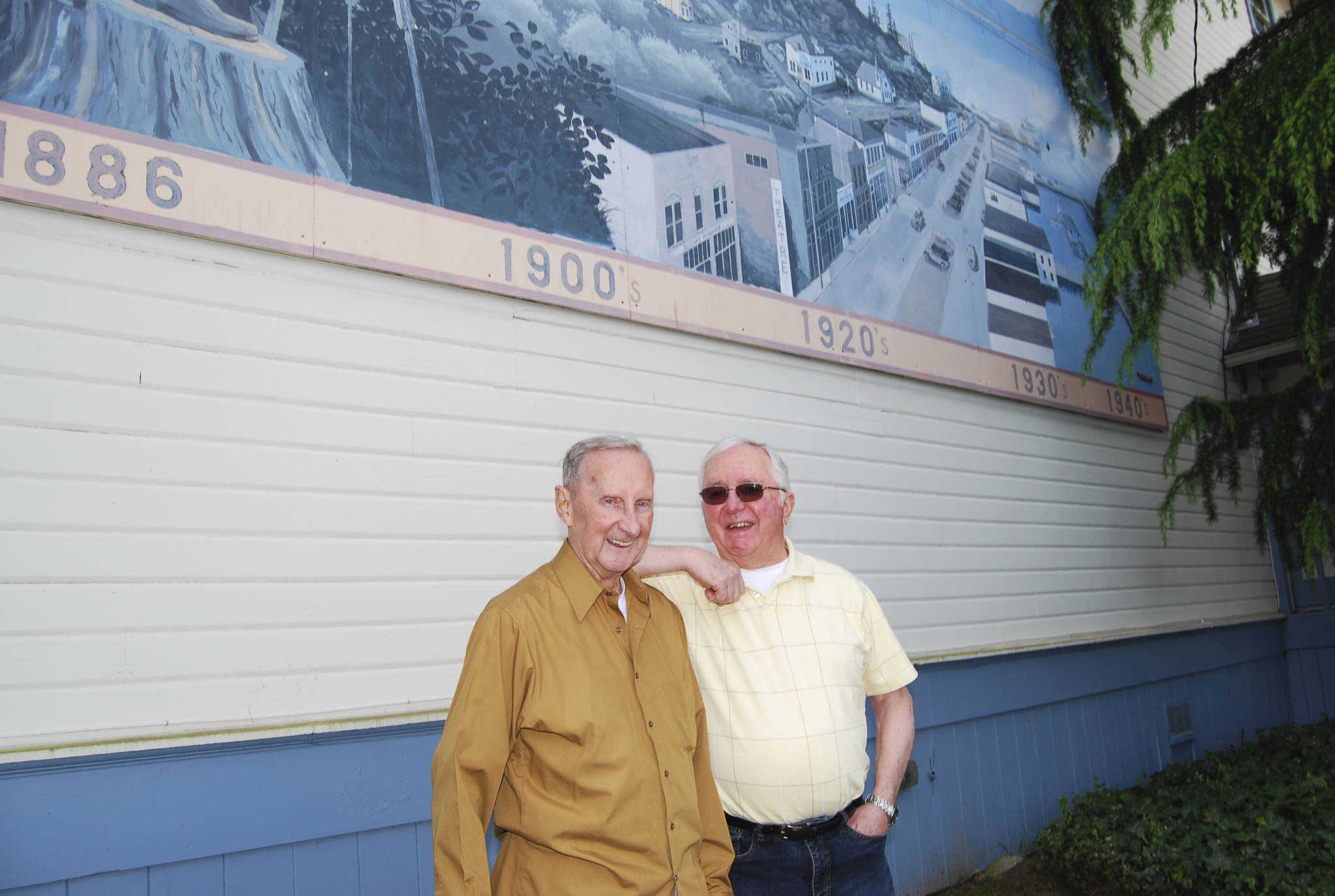 Bob Ulsh (left) and Roy Carr stand in front of a 12-panel mural outside the Sidney Art Gallery and Museum that Ulsh painted depicting a timeline of Port Orchard’s history beginning in the 1880s. (Bob Smith | Kitsap Daily News photo 2016)