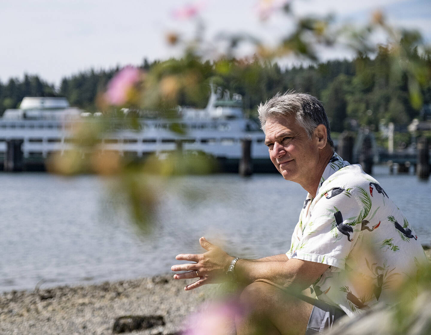Greg Sugden, in his Hawaiian shirt, relaxes on a sandy beach on Bainbridge Island, with one of the state ferries in the background. Damon Williams/Kitsap News Group