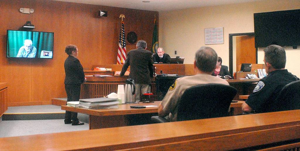 Clinton R. Caldwell, 67, of Sequim appears on a television monitor in Clallam County Superior Court on Tuesday as Chief Criminal Deputy Prosecuting Attorney Michele Devlin and attorney John Hayden stand before Superior Court Judge Chris Melly in Port Angeles. Witnessing the proceeding from the prosecutors table were Clallam County Sheriff William Benedict and Port Angeles police Chief Brian Smith. (Keith Thorpe/Peninsula Daily News)