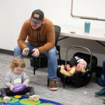 Dad watches his kid play with a drum pad. Even the child in the car seat is enjoying the performance.