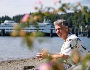 Greg Sugden, in his Hawaiian shirt, relaxes on a sandy beach on Bainbridge Island, with one of the state ferries in the background. Damon Williams/Kitsap News Group